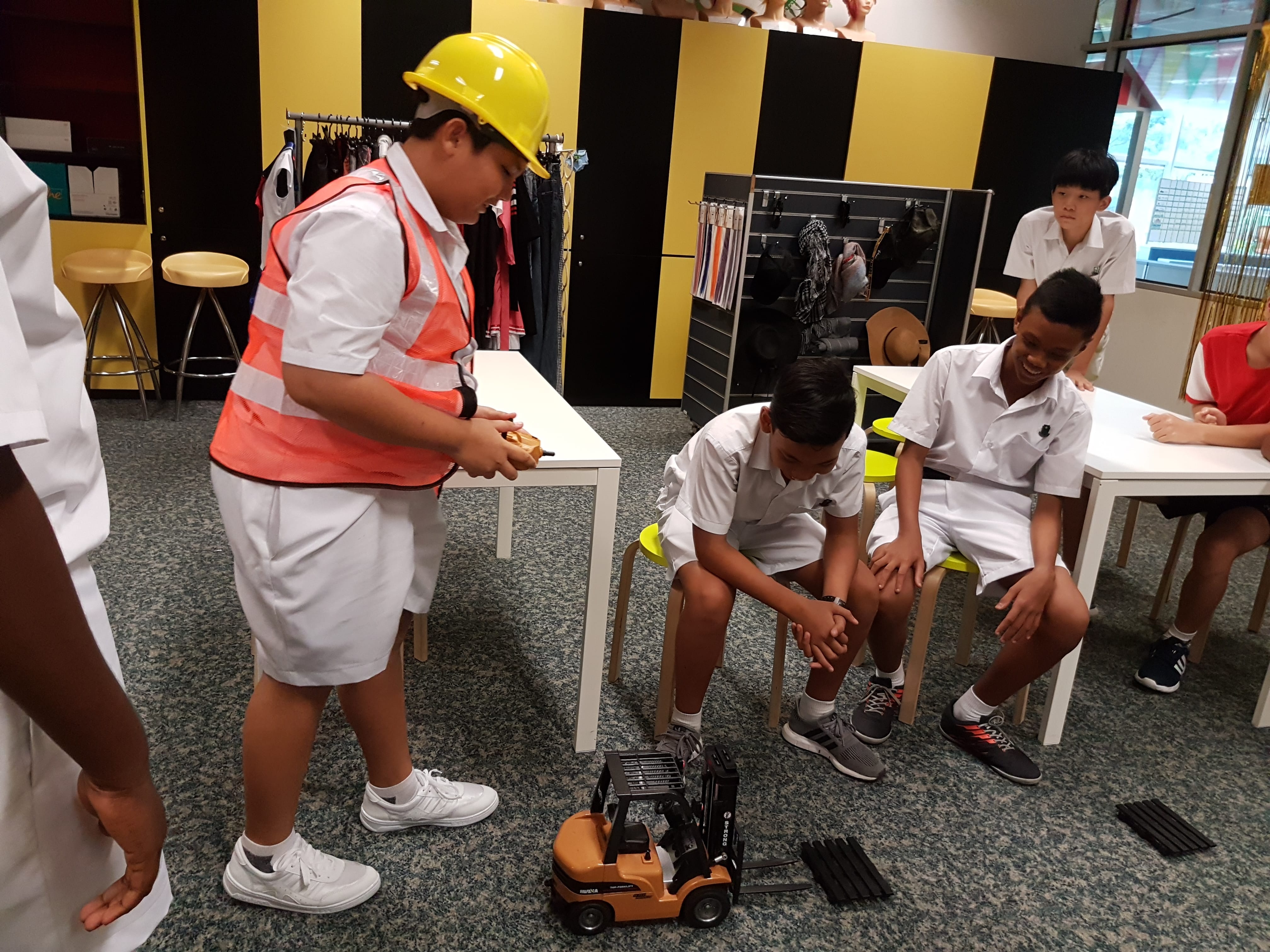 Boy in hard hat and safety vest uses a toy forklift as classmates in uniforms watch and laugh.